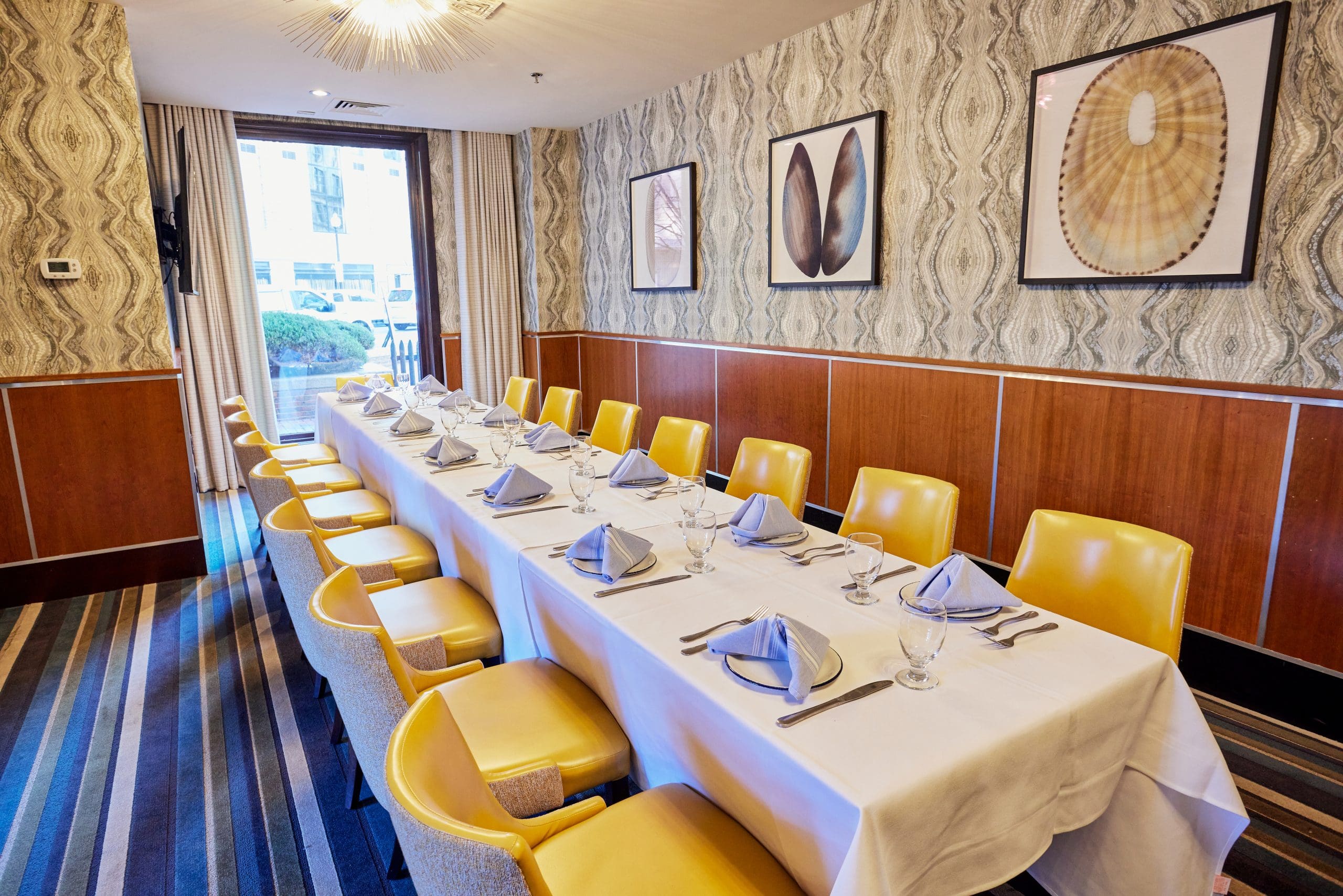 A long dining table with a white tablecloth is set with folded gray napkins, glassware, and silverware in an elegant room. Yellow chairs line both sides of the table. The walls have patterned wallpaper and are adorned with framed art. A high window offers natural light.