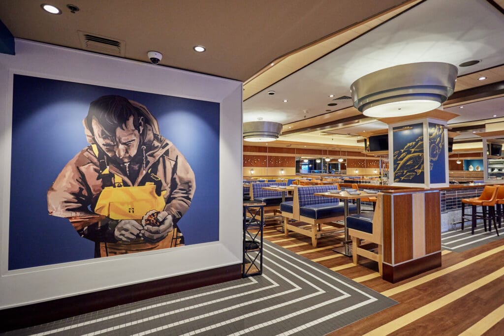 A modern restaurant interior featuring wooden and blue upholstered seating. The flooring has various designs including wood and patterned tiles. A large mural of a fisherman in yellow overalls is prominently displayed on one wall.