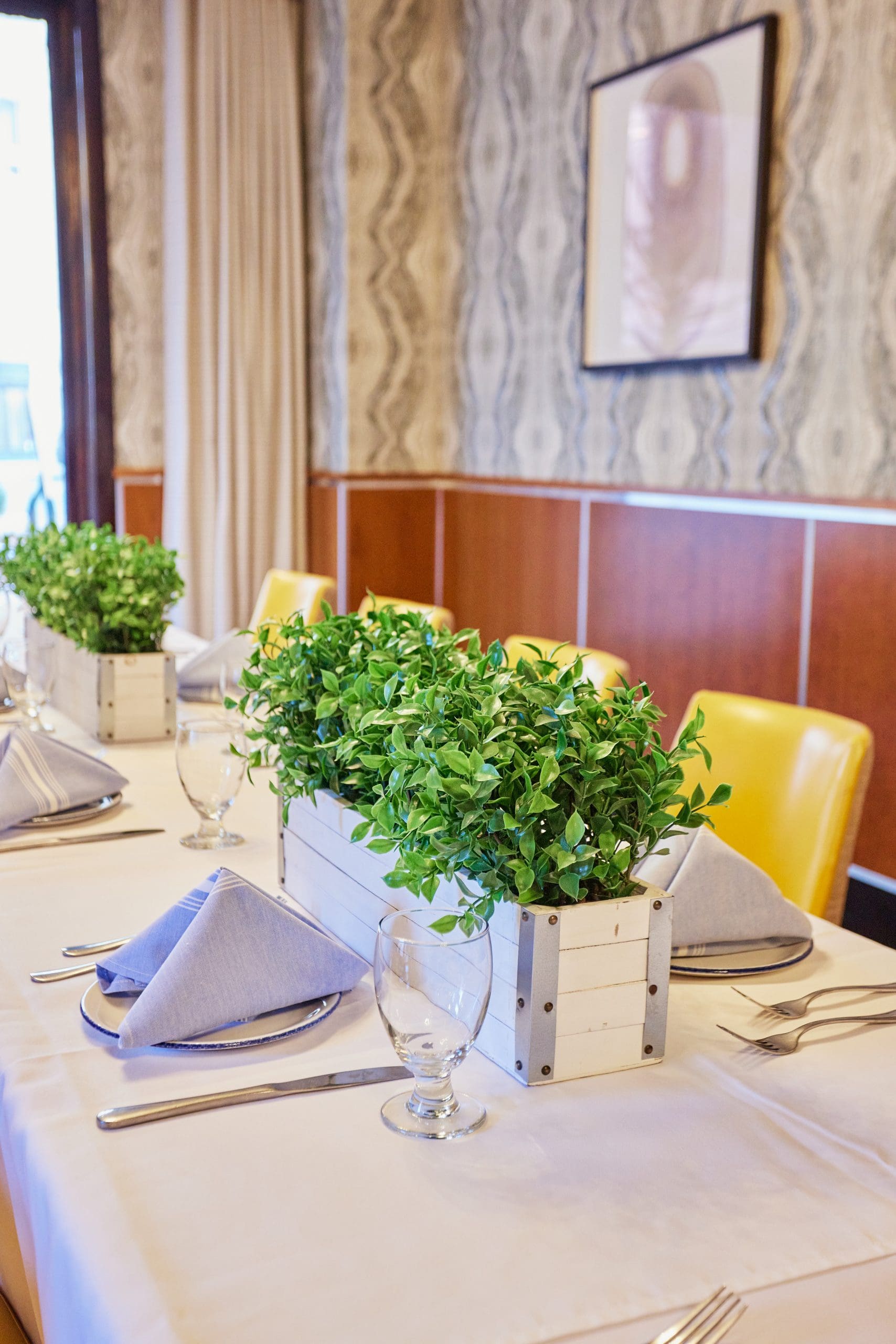 A stylish dining setup featuring a long table with a white tablecloth, set with neatly folded gray napkins, glassware, and cutlery. Wooden planters holding green plants serve as centerpieces. Yellow chairs line the table, and abstract art decorates the wall in the background.