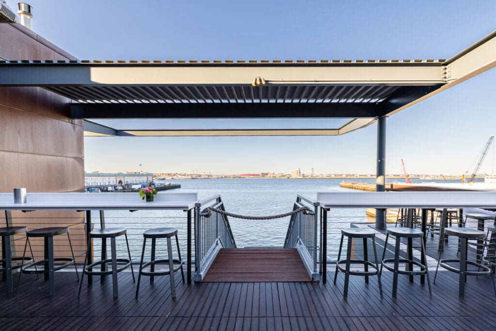 A waterfront seating area with high tables and stools under a pergola. The area overlooks a calm body of water, with a clear blue sky and various industrial buildings and cranes visible in the distance. A floral arrangement sits on one of the tables.