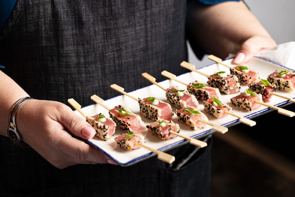 Person holding a rectangular platter with skewers of seared tuna garnished with green onions and sesame seeds. The person is wearing a dark apron and blue shirt. The platter features neatly arranged skewers, each with several pieces of tuna.