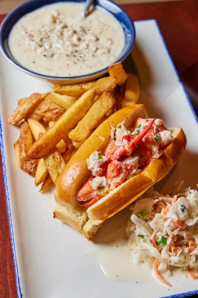 Enjoy the Express Lunch Combo from Legal Sea Foods: a lobster roll in a toasted bun, thick-cut fries, creamy coleslaw, and a bowl of clam chowder with a spoon—all on one plate for a satisfying midday meal.