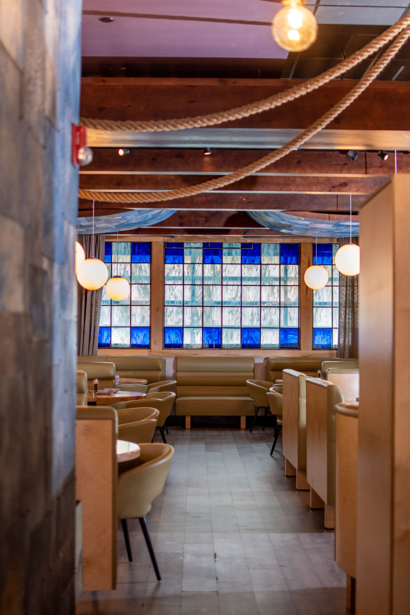 A cozy restaurant interior with beige leather booths and wooden tables. The ceiling features exposed wooden beams, hanging bulbs, and decorative ropes. Blue and white mosaic windows at the far end provide ambient lighting. The floor is tiled.