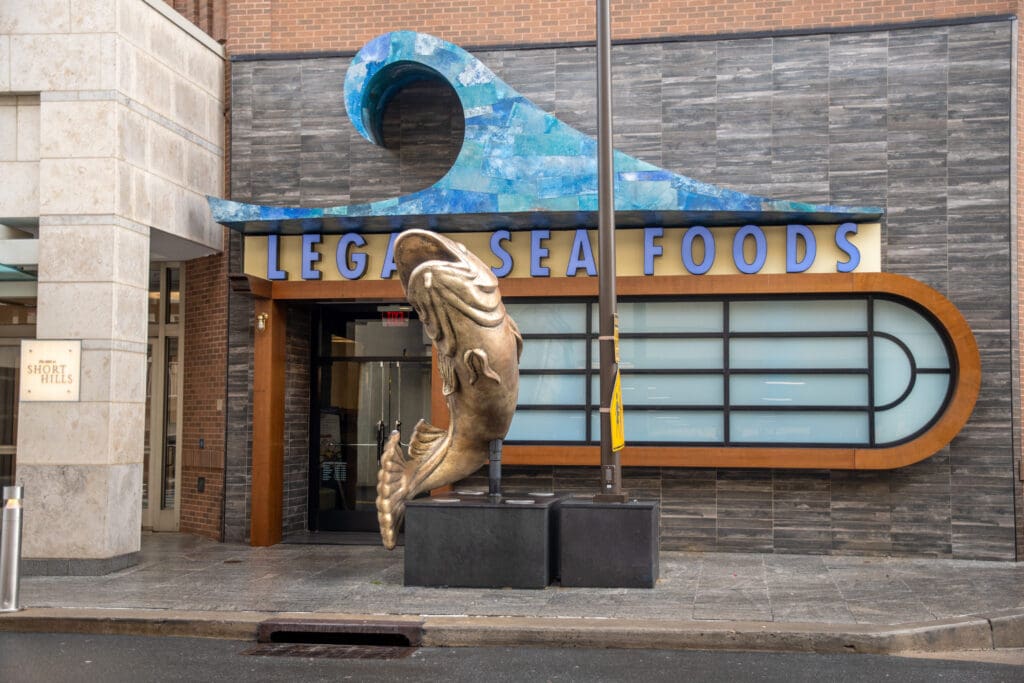 The storefront of Legal Sea Foods with a large fish sculpture emerging through the sidewalk. Above the entrance is a sign with a blue wave design. The building features brick and stonework with large windows. A sign for 