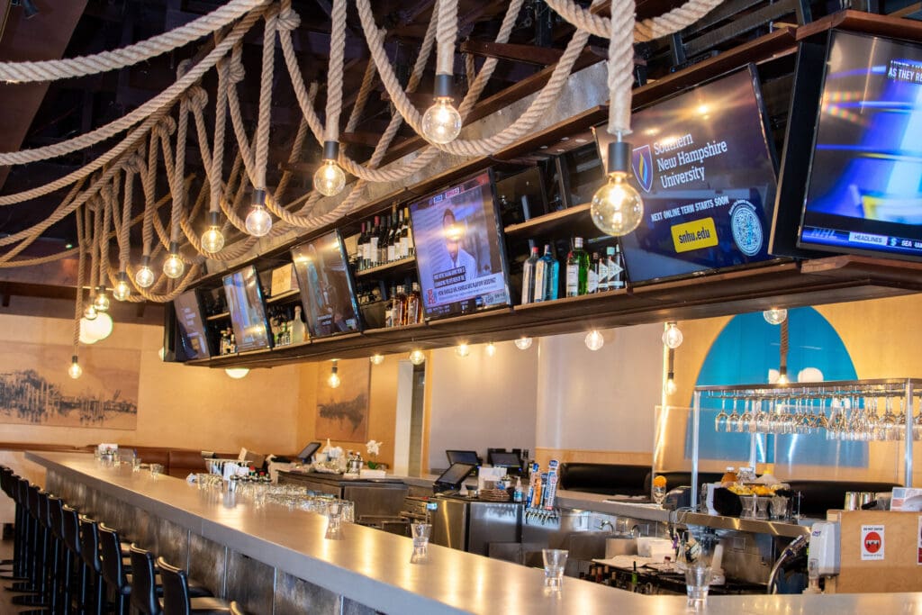 A modern bar with a long counter, high stools, hanging light bulbs, and multiple flat-screen TVs displaying various channels. The decor includes thick ropes hanging from the ceiling. Shelves behind the bar hold bottles, glasses, and other barware.
