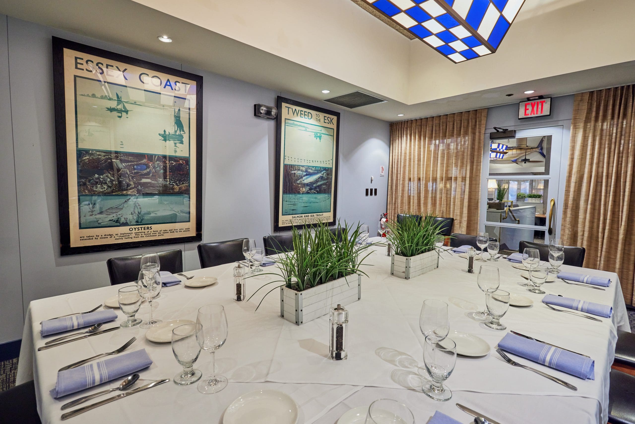 A dining room with a long, white table set with plates, glasses, and blue napkins. The table is decorated with potted plants. The walls have vintage travel posters, and there is an exit sign above a door with curtains adjacent to it.