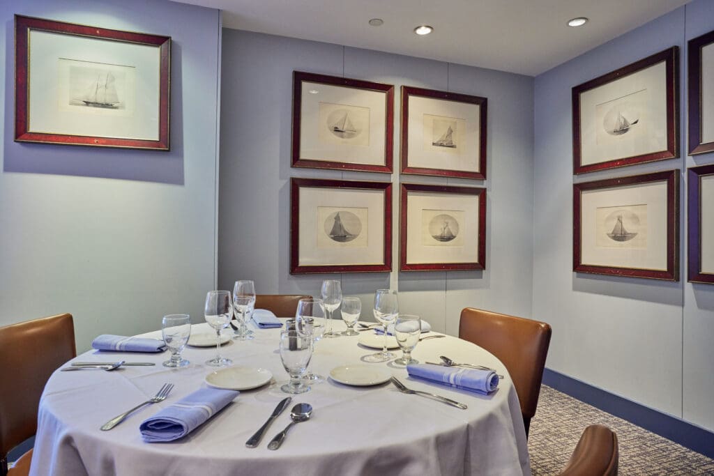 A round dining table set for six with white linens, glasses, and silverware. Brown chairs surround the table. The light blue walls display framed black and white drawings of boats. Recessed lighting illuminates the room, creating a cozy ambiance.