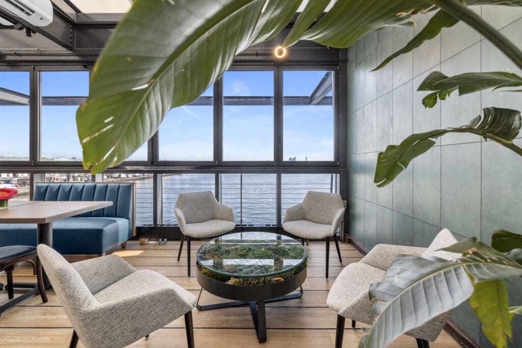 A modern indoor lounge area with large windows offering a scenic water view. The space features four gray cushioned chairs around a glass-top coffee table with plants inside it. Green walls and leafy plants add a touch of nature to the stylish décor.