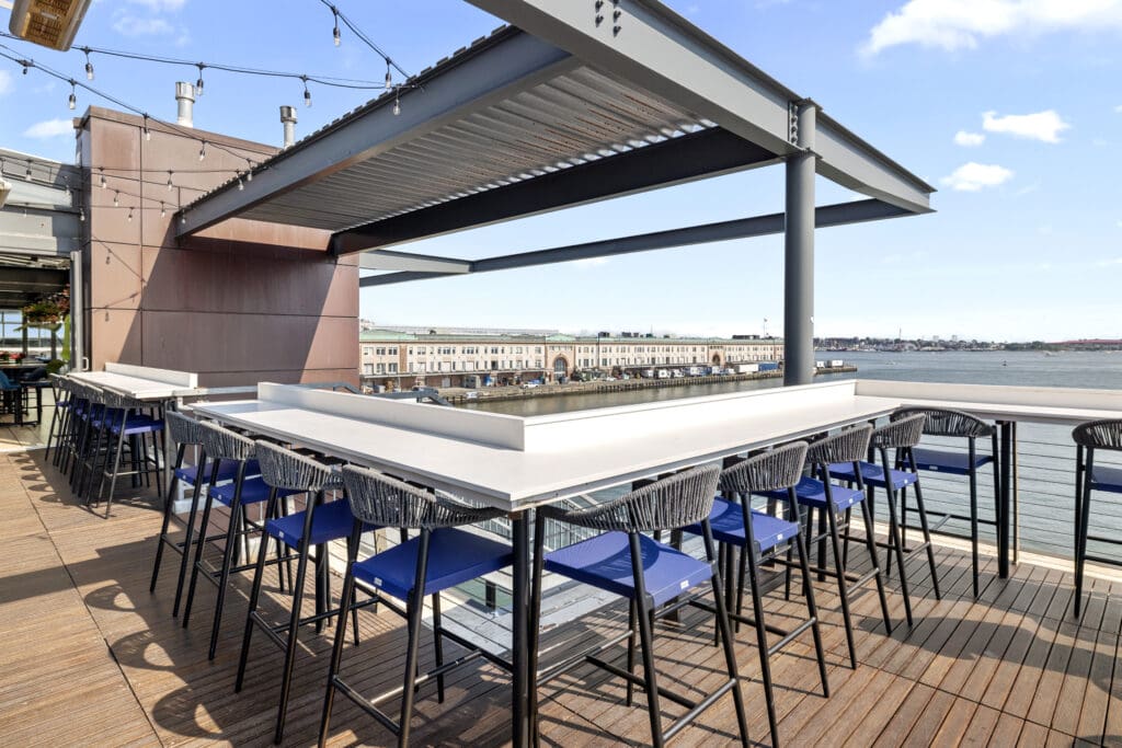 A rooftop patio with long, high tables and blue bar stools overlooks a waterfront. The patio is shaded by a modern pergola. String lights are draped above, adding to the ambiance. In the background, buildings and a body of water are visible under a blue sky.