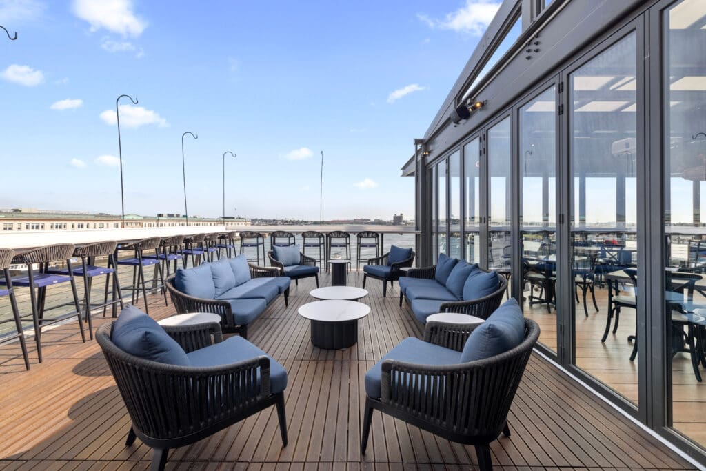 A rooftop terrace features several blue cushioned seats arranged around circular tables, with a modern glass building wall on one side. The terrace overlooks a cityscape with a clear blue sky in the background.
