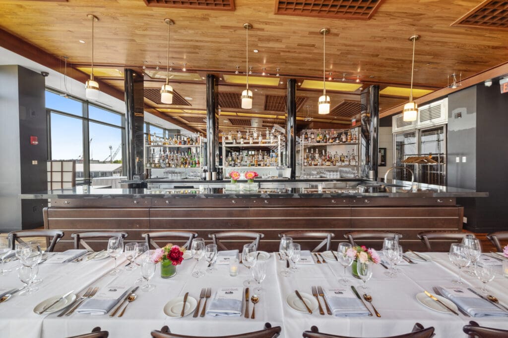 A stylish restaurant with a long bar area, decorated with hanging lights and stocked shelves of bottles. In the foreground, a table is set elegantly with white tablecloth, silverware, glassware, and small floral centerpieces. Wood-paneled ceiling adds warmth.