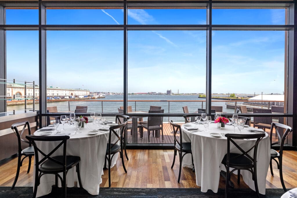 Elegant restaurant dining area with round tables covered in white tablecloths and set for a meal. Large floor-to-ceiling windows offer a picturesque view of a calm waterfront and clear blue sky. Dark wooden flooring and chairs complete the sophisticated ambiance.