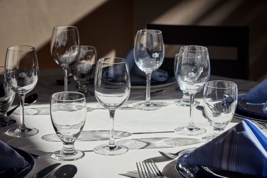 A dining table is elegantly set with various glassware, including wine and water glasses. The table is adorned with blue napkins folded neatly, accompanied by silverware. Shadows cast across the table enhance the sophisticated ambiance.