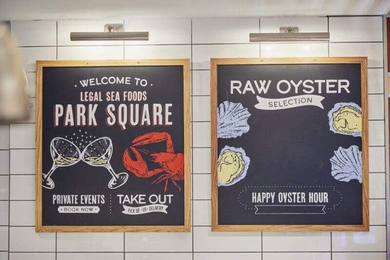Two chalkboard-style signs in light wooden frames on a white tiled wall. The left sign advertises “Legal Sea Foods Park Square” with images of a lobster and clams. The right sign reads “Raw Oyster Selection” with illustrations of oysters and mentions “Happy Oyster Hour.”.