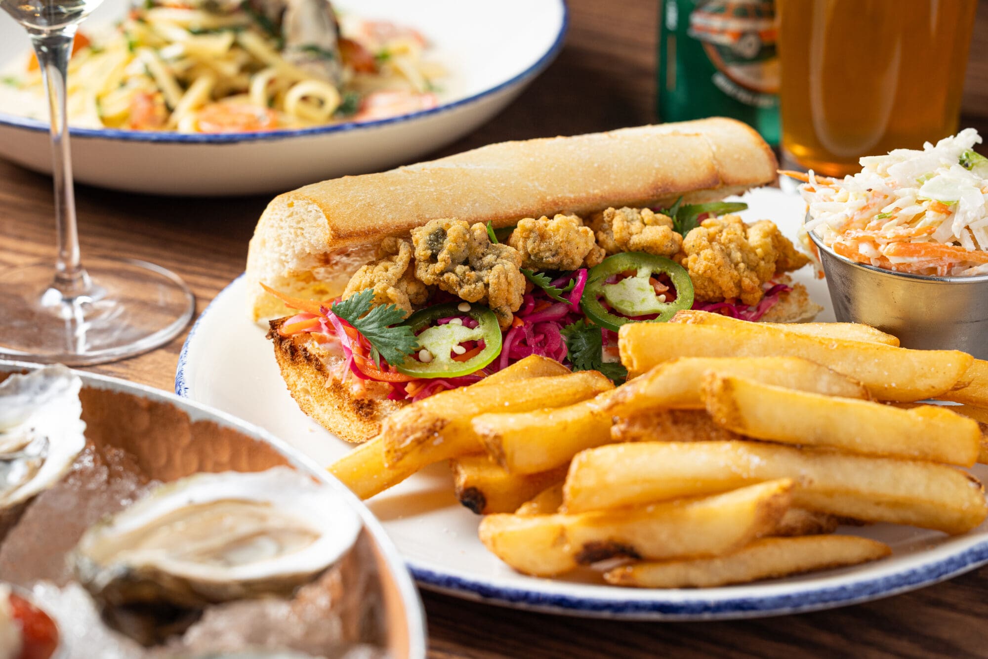 A plate with a fried oyster po’ boy sandwich topped with jalapeños, red cabbage, and cilantro, served with thick-cut fries and a side of coleslaw. Pasta and drinks are visible in the background.