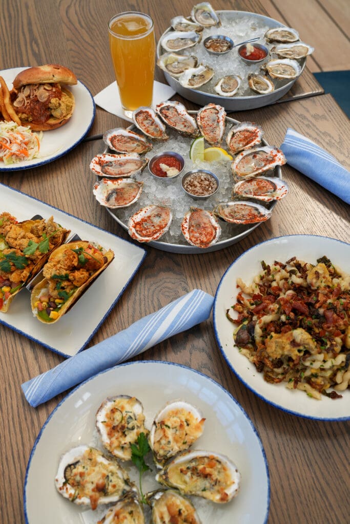 Plates of oysters, tacos, a sandwich with fries, a vegetable dish, and a glass of beer are arranged on a wooden table with blue napkins.