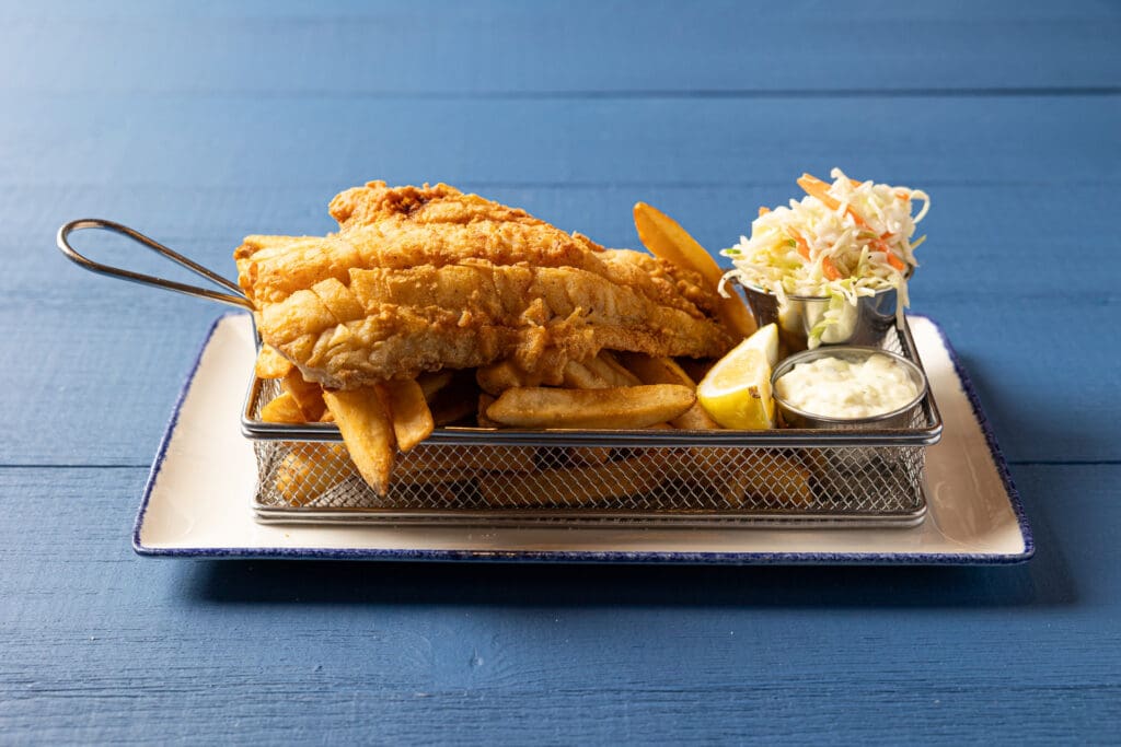 A serving of fish and chips is presented on a white plate with a blue rim. The meal includes battered fish and fries in a metal basket, a lemon wedge, a small dish of tartar sauce, and a side of coleslaw. The background is a blue surface.
