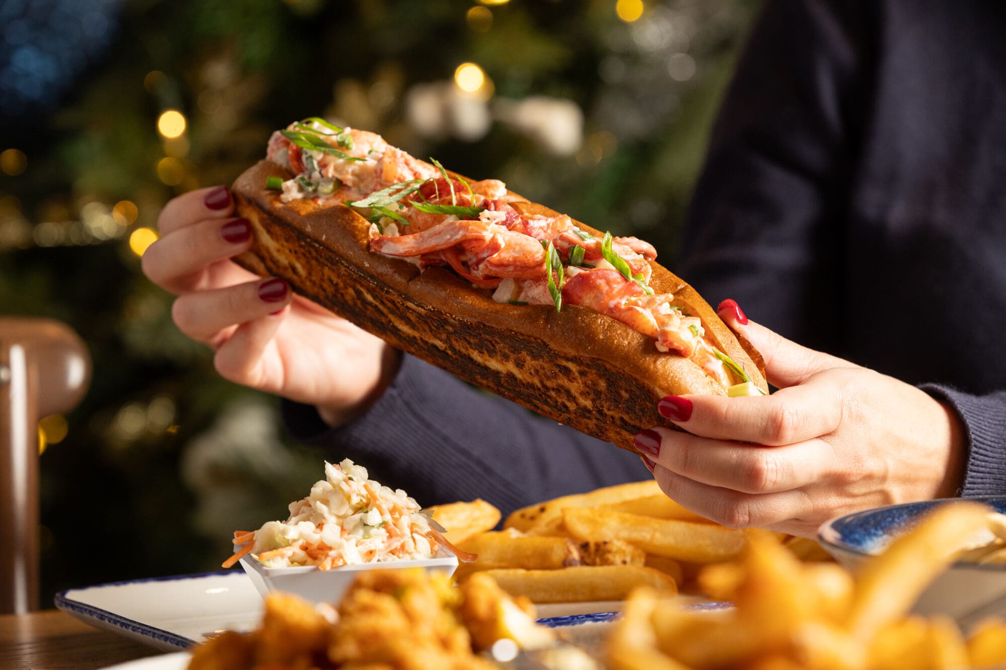 A person holding a toasted lobster roll garnished with herbs, with a plate of French fries, coleslaw, and fried food visible on the table in the foreground.