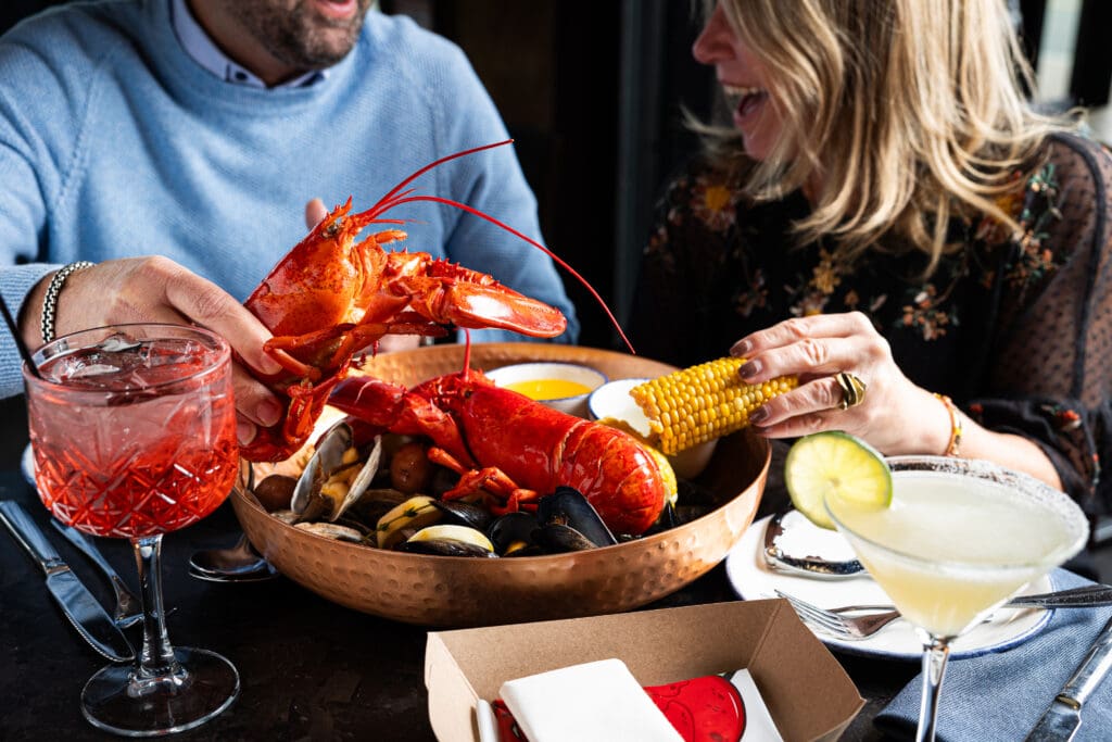 A couple enjoys a seafood feast at a restaurant, featuring a large bowl with a cooked lobster, mussels, and corn on the cob. They are laughing and holding food. Two cocktails are on the table: a pink drink and a margarita with a lime slice.