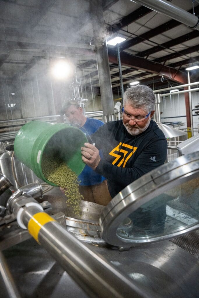 A man pours a bucket of green hops into a large brewing tank in an industrial brewery, while another man stands behind him amid steam and stainless steel equipment.