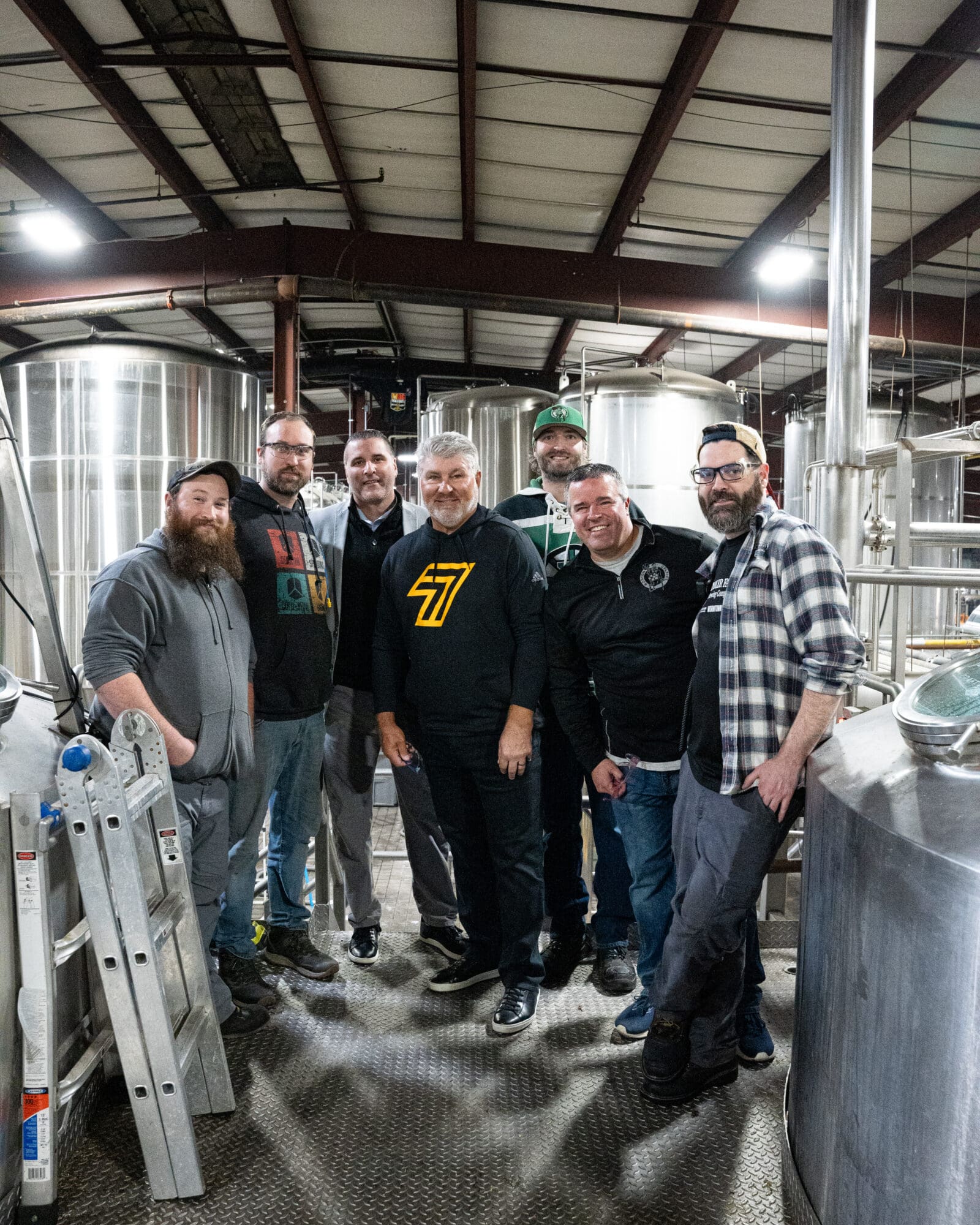 Seven people stand smiling in a brewery surrounded by large metal tanks. A ladder leans on the left. They wear casual clothing, including hoodies, jackets, and hats. The ceiling is supported by metal beams.