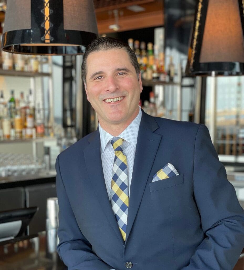 A smiling man in a suit and tie stands in front of a bar shelf with various bottles. He wears a pocket square that matches his tie. The background has large pendant lights and a modern bar interior.