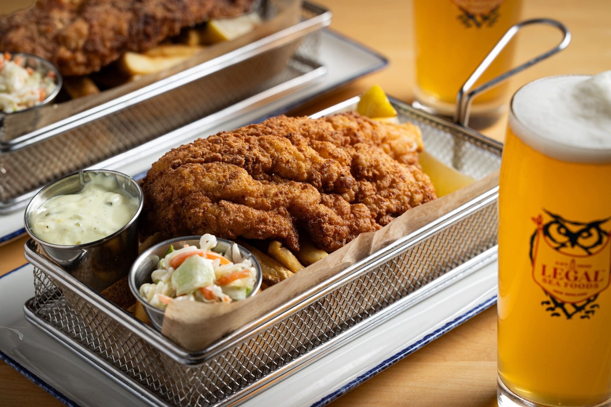 Close-up of a basket with fried fish, golden fries, tartar sauce, and coleslaw. A tall glass of beer is nearby, with part of another identical meal in the background, both on wooden tables. The logo on the glass reads 