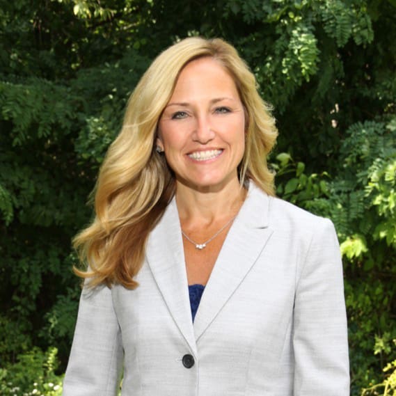 A woman with long blond hair smiles while standing outdoors against a backdrop of green foliage. She is wearing a light gray blazer and a blue top.