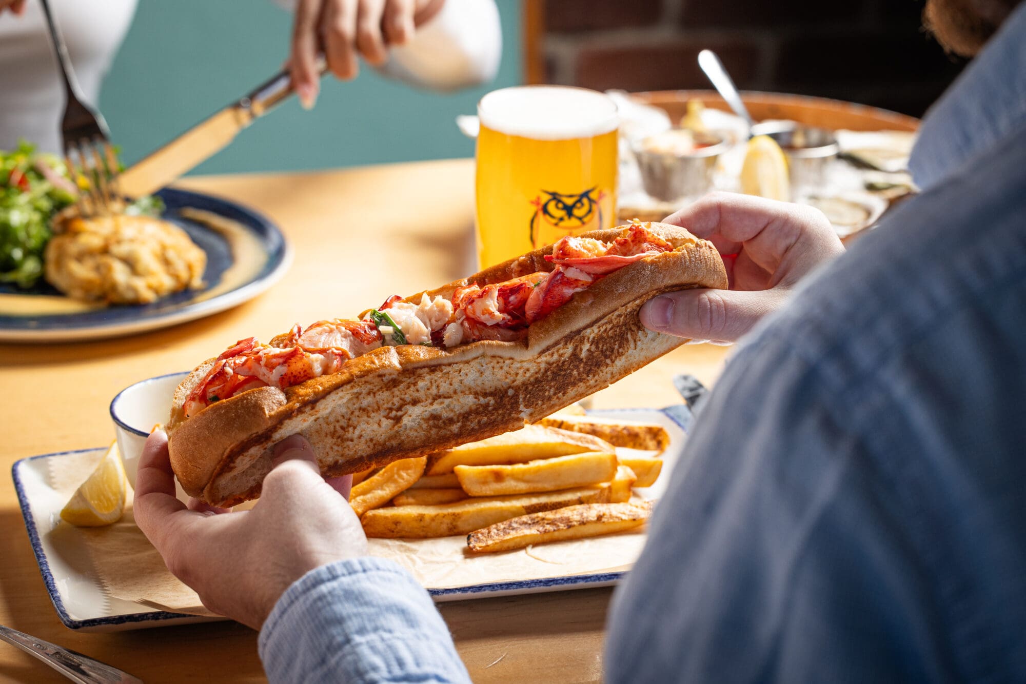 A person holding a lobster roll on a plate with fries. Another person is seen in the background eating a salad and an omelet. A glass of beer is on the table, alongside bowls with various condiments.
