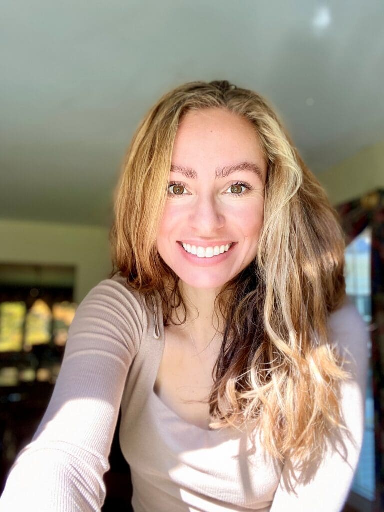 A woman with long, wavy hair and a big smile is taking a selfie in a well-lit room. She is wearing a beige top and the background shows a window with sunlight streaming in.