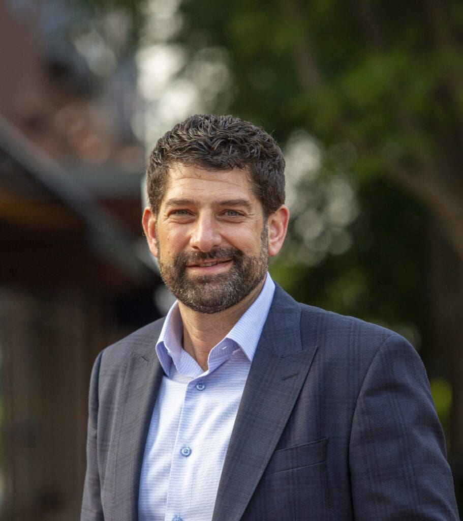 A man with curly dark hair and a beard is smiling, wearing a blue collared shirt and a gray blazer. The background is blurred with greenery and a hint of a building.
