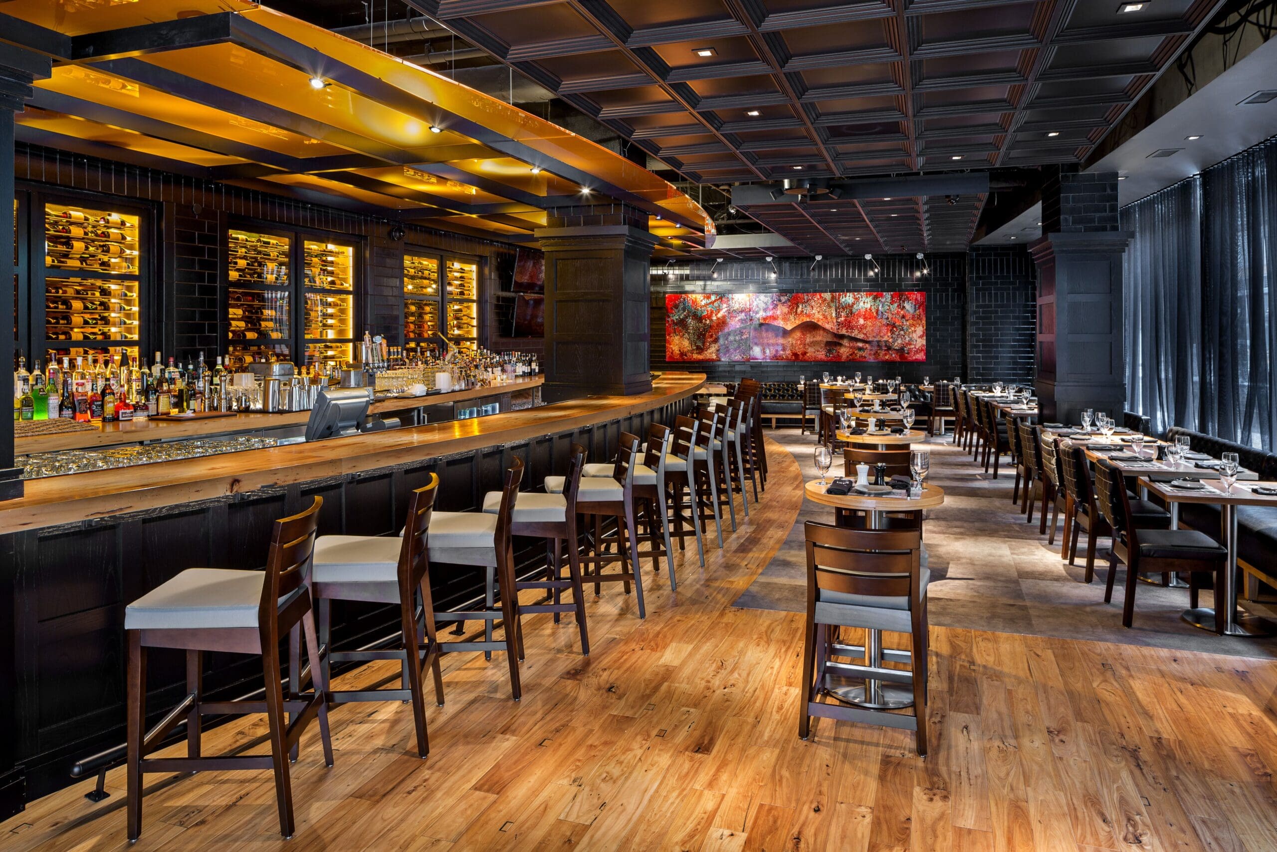Modern restaurant interior with a long wooden bar on the left, high-backed chairs, and a well-stocked shelf of bottles. Dining tables are set neatly on the right under warm lighting, with a colorful mural on the back wall.