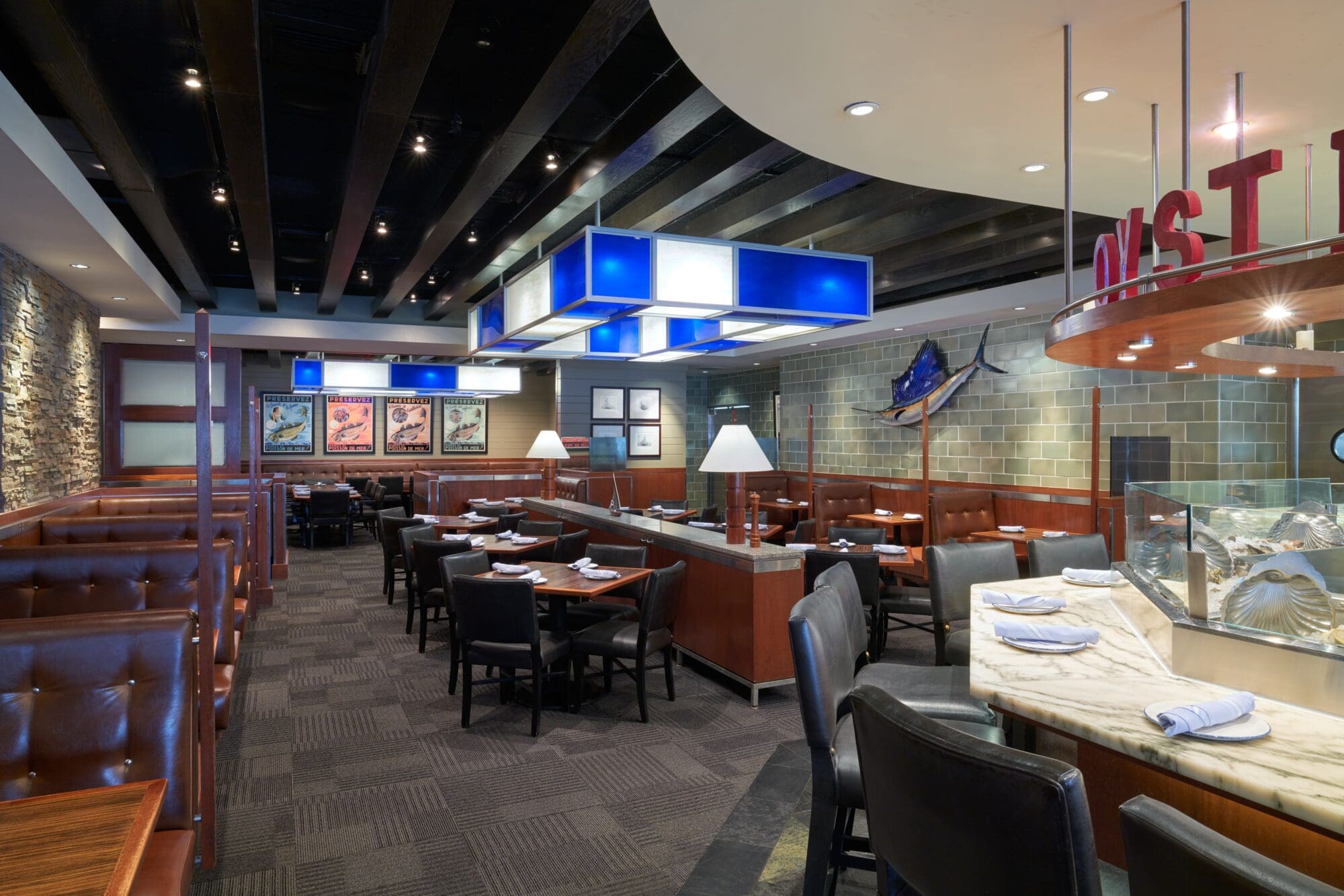 Interior of a modern restaurant with brown leather booths, dark wood accents, and a marble-topped bar. Soft lighting, framed art on the walls, and a decorative blue fish sculpture create a cozy atmosphere. Tables are set with white napkins.