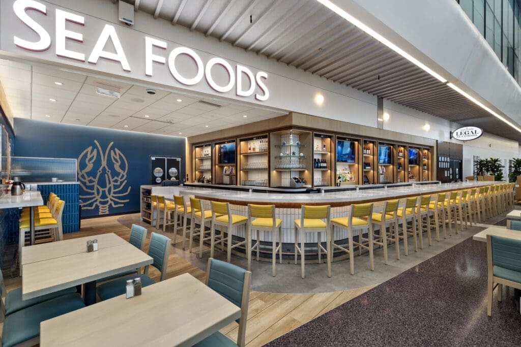 A modern seafood restaurant with a large, curved bar surrounded by yellow chairs. The wall displays shelves of glassware and bottles.