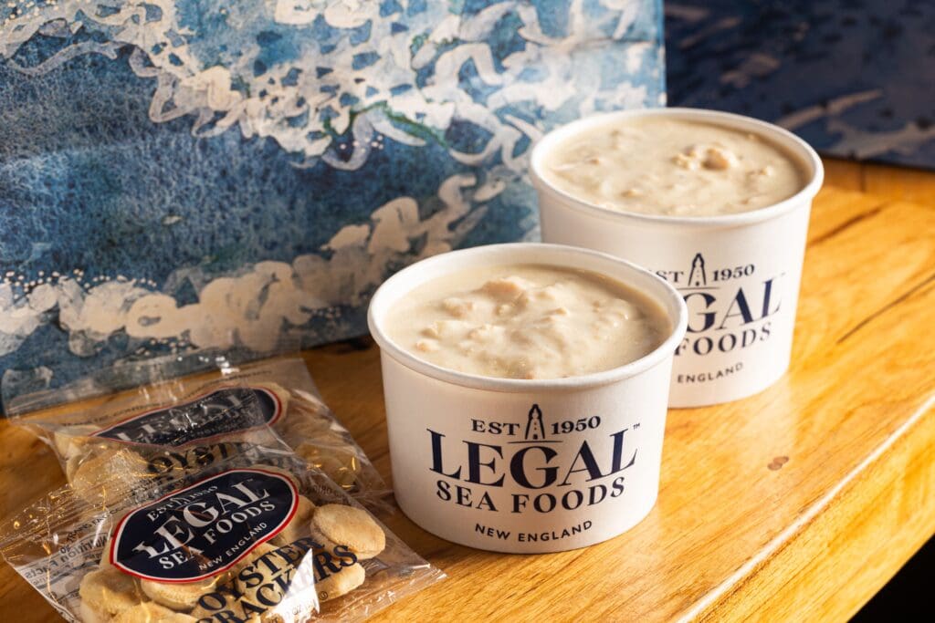 Two cups of clam chowder from Legal Sea Foods placed on a wooden surface. Each cup is branded with the restaurant's logo and is accompanied by a small pack of oyster crackers. A textured blue and white background is visible.
