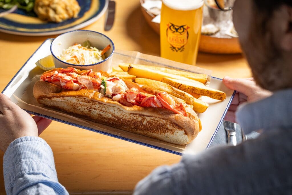 A person holds a plate with a lobster roll topped with pieces of lobster and garnished with herbs, alongside thick-cut fries. A side of coleslaw and a lemon wedge accompany the dish. In the background, there's a glass with a logo and other plates.