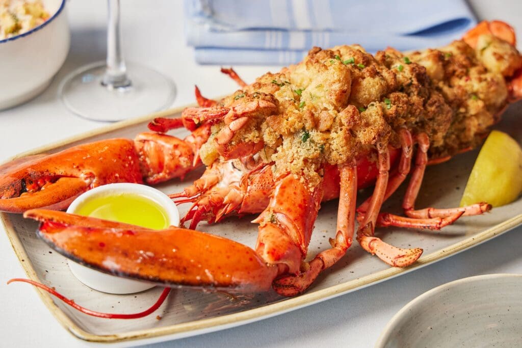 Stuffed lobster on a rectangular plate, garnished with breadcrumbs and herbs. A small dish of melted butter and a lemon wedge are on the side. A glass and a folded napkin are visible in the background.