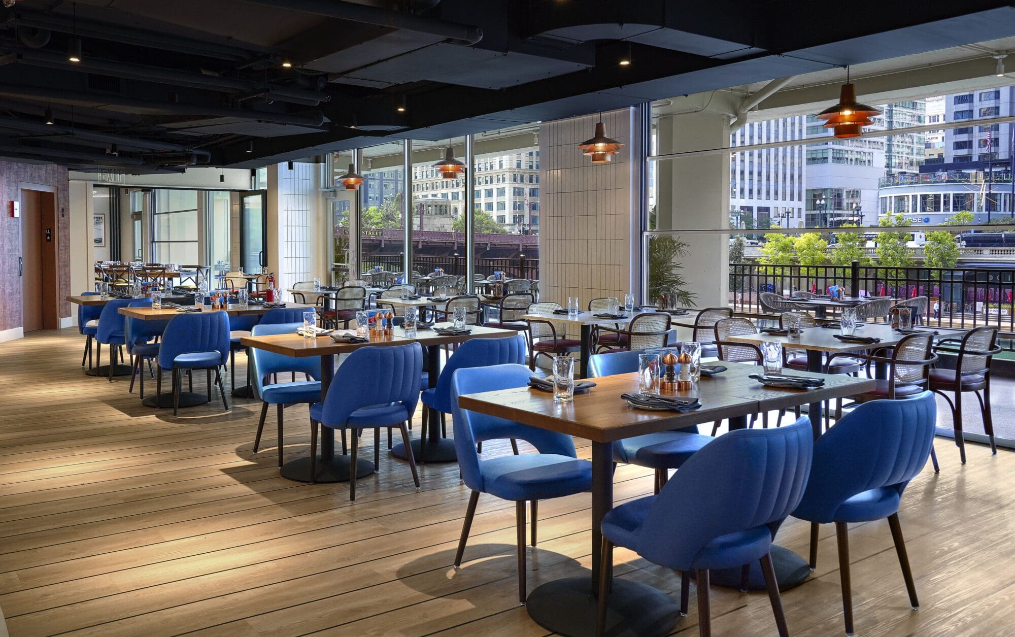 Spacious restaurant interior with wooden floors, featuring several tables set for dining. Each table has blue chairs, cutlery, and condiments. Large windows offer a view of urban buildings and greenery outside. Warm, inviting atmosphere.