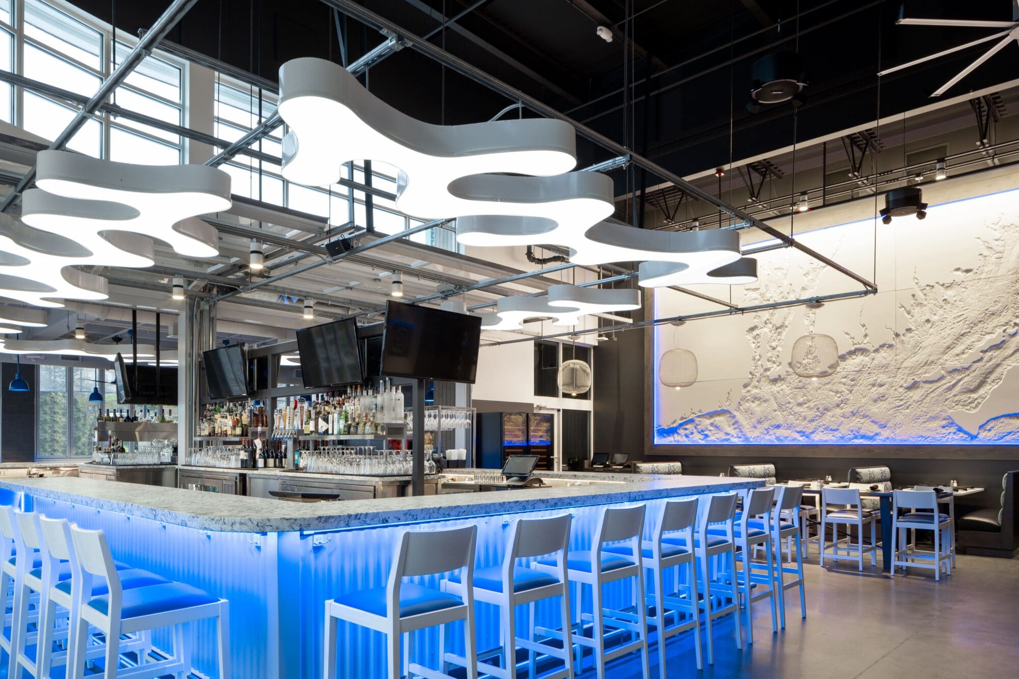 Modern bar interior with a futuristic design. The bar counter is illuminated with blue lighting and surrounded by white stools. Abstract white ceiling lights hang above. Multiple flat-screen TVs are mounted around the bar, with a textured wall in the background.