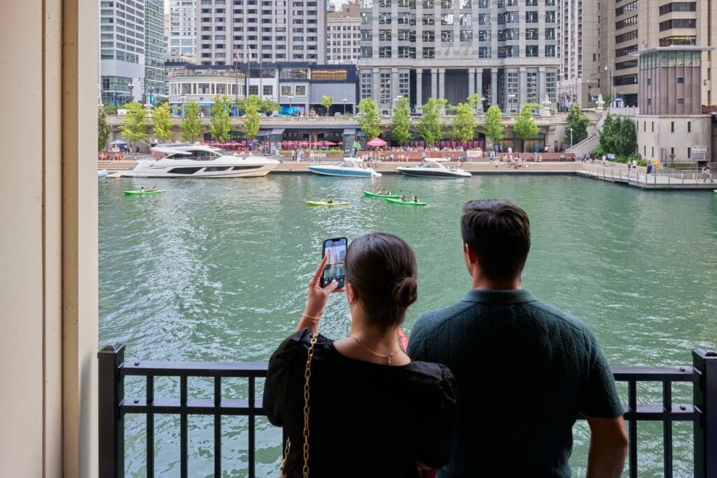 A man and woman stand by a railing, looking at a river with motorboats and kayakers. The woman holds up her phone to take a photo. In the background, there are buildings and a riverside park with people walking.