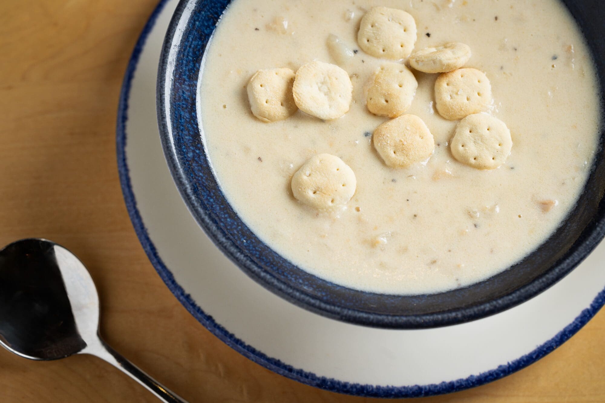 A bowl of creamy clam chowder topped with round oyster crackers is set on a wooden table. A metal spoon rests on the table beside the bowl. The chowder is served in a dark blue bowl placed on a white plate.