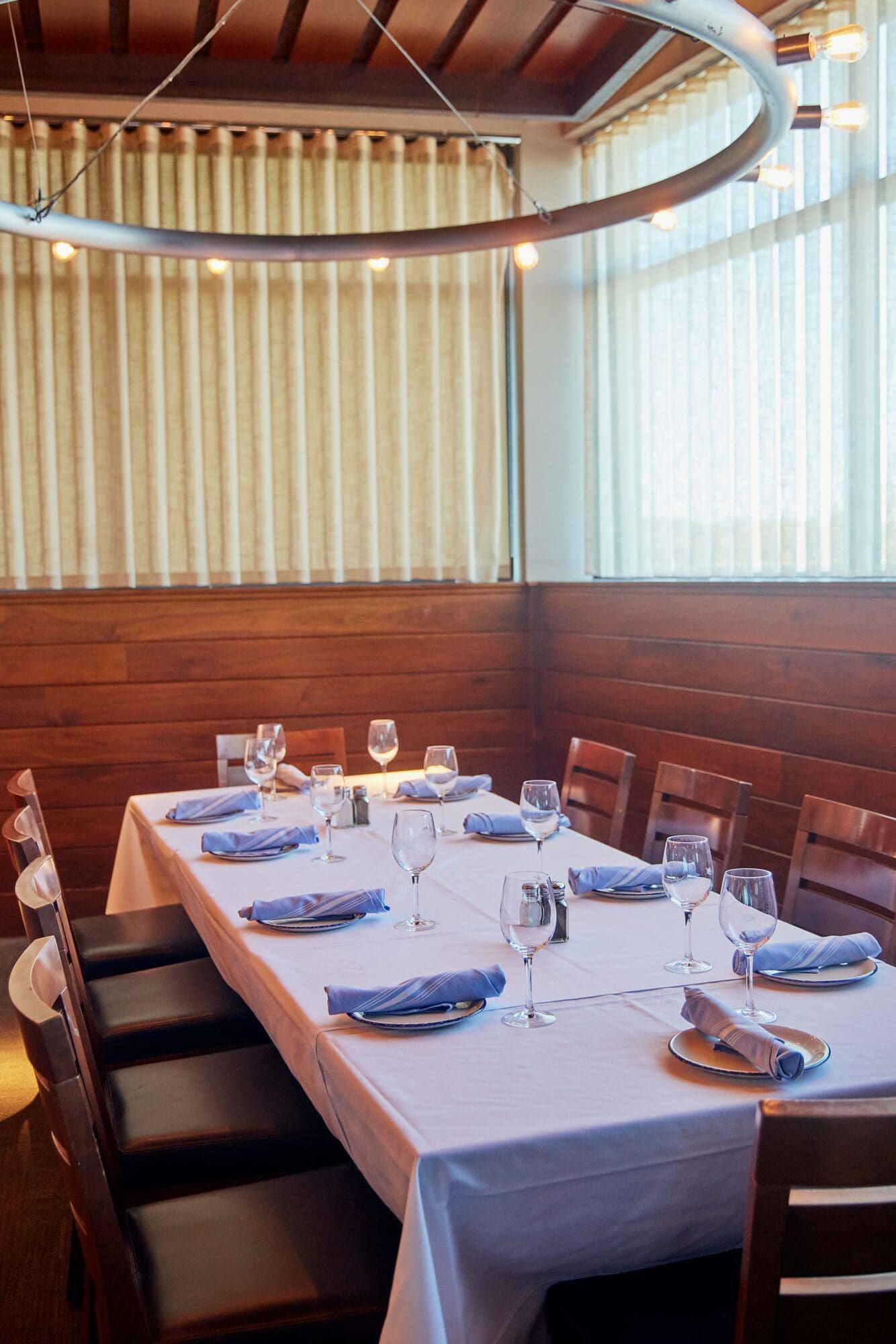 A dining table set for eight with a white tablecloth, blue napkins, and empty wine glasses in a warmly lit room. The room has wood-paneled walls, tall windows with light-colored curtains, and a circular light fixture above.