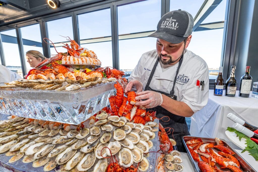 A chef arranges a display of seafood on an ice sculpture, featuring oysters, prawns, and lobster tails. Bottles of wine are visible in the background. The chef wears gloves and a hat, focused on the arrangement.