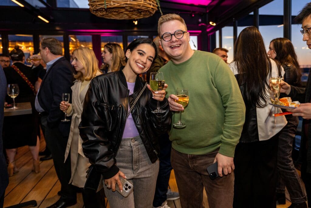 Two people smiling and posing together at a lively indoor social event, each holding a glass of white wine, with other guests mingling in the background.