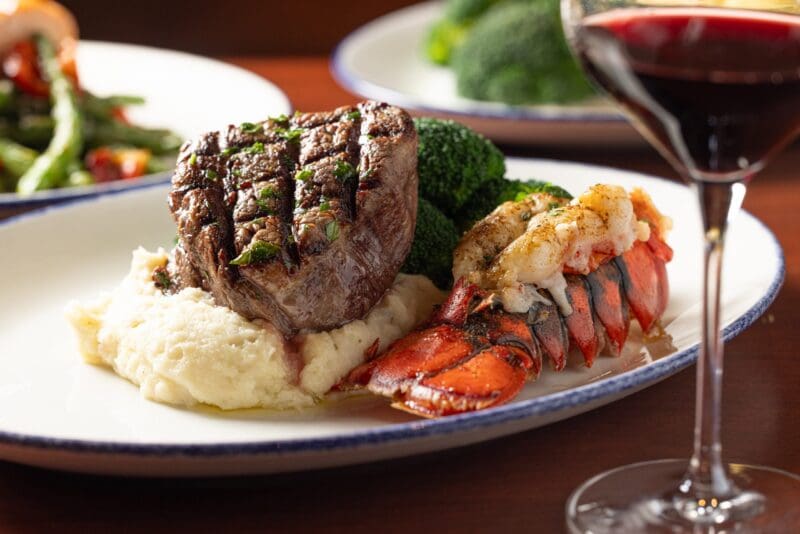 A plate with grilled steak on mashed potatoes, a lobster tail, and steamed broccoli, with a glass of red wine in the foreground and vegetables in the background.