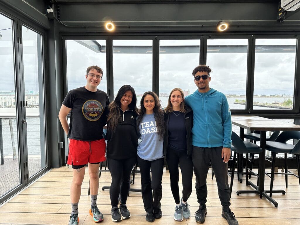 Five people stand side by side smiling indoors on a wooden floor, with tall windows showing a cloudy sky and water outside. One person wears a 