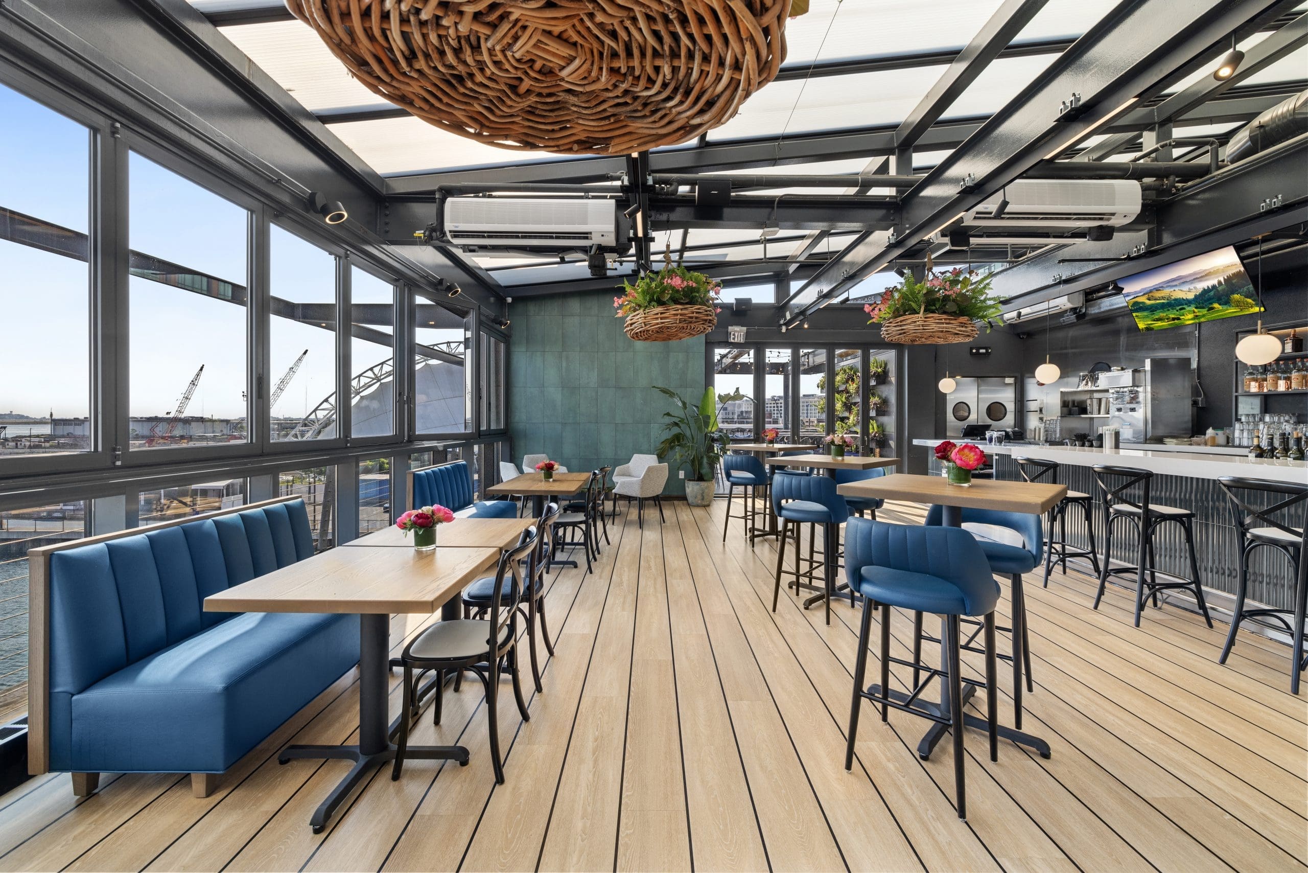 Modern rooftop restaurant with wood flooring, blue seating, hanging plants, large windows showing an industrial waterfront view, and a bar on the right side with stools and a TV screen.