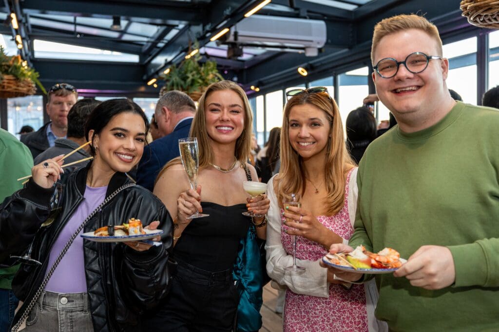 Four people smile at the camera while holding plates of sushi and drinks at an indoor social gathering. The group appears happy, with one person holding chopsticks and others raising glasses in a toast.