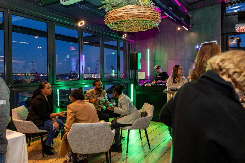 A group of people are socializing in a modern, neon-lit rooftop lounge with large windows showing a cityscape at dusk. A DJ is playing music near the bar, and guests are seated and mingling around tables.