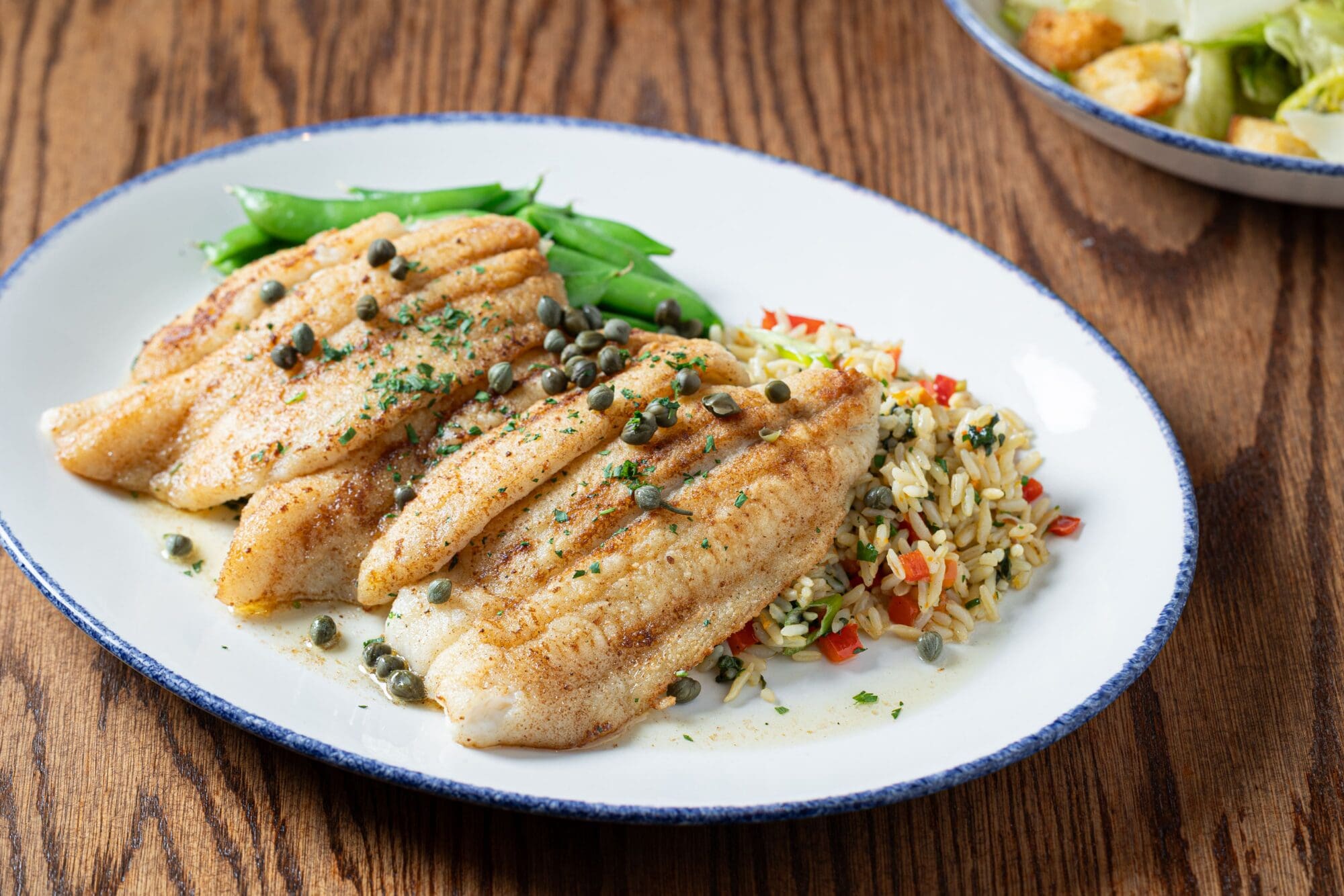 Grilled fish fillets topped with capers, served with green beans and vegetable rice on a white oval plate, placed on a wooden table.